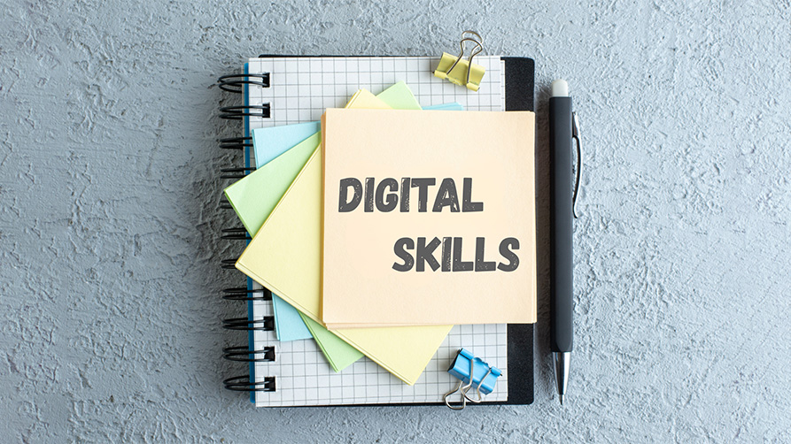 Vue de dessus d'un carnet à spirales posé sur une surface texturée grise, entouré de trombones colorés et d'un stylo noir. Sur le carnet, une pile de notes adhésives colorées (jaune, vert, bleu) avec une note de tête orange portant l'inscription textuelle "DIGITAL SKILLS" en lettres capitales grises