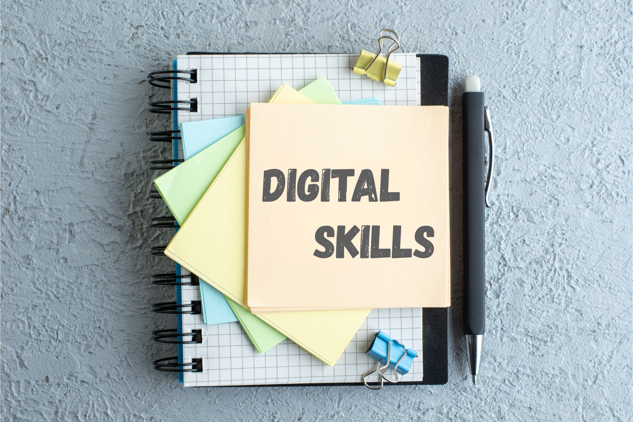 Vue de dessus d'un carnet à spirales posé sur une surface texturée grise, entouré de trombones colorés et d'un stylo noir. Sur le carnet, une pile de notes adhésives colorées (jaune, vert, bleu) avec une note de tête orange portant l'inscription textuelle "DIGITAL SKILLS" en lettres capitales grises