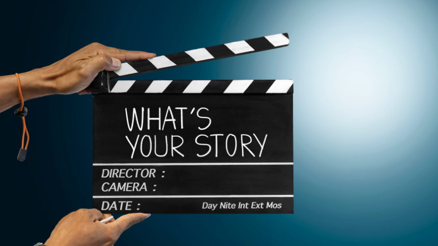 Des mains tiennent un clap de cinéma noir (clapperboard) sur fond bleu, sur lequel est écrit à la craie la question "WHAT'S YOUR STORY" (Quelle est votre histoire).