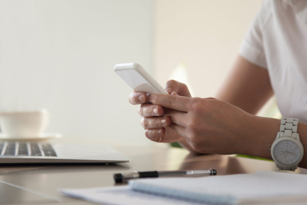 Gros plan sur les mains d'une femme professionnelle lisant un message sur un smartphone blanc à son bureau. Illustration de l'immédiateté et du fort taux d'ouverture des campagnes de SMS marketing.